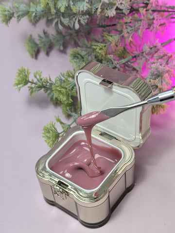 Lemmy Builder Gel 23 - Mocca Mousse in an open container with application tool, surrounded by foliage, showcasing creamy texture.